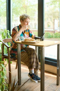 Myoelektrik kollu genç bir bayan akıllı telefon kullanıyor ve kafede otururken kahvaltıda sandviçle kahve içiyor.