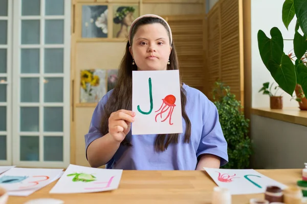 年轻而严肃的老师坐在电脑前或照相机前的桌子旁 展示着画有绿色字母J和红色水母的纸 — 图库照片