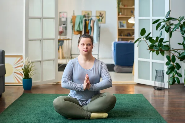 Young woman with disability sitting in pose of lotus on the floor of spacious living room and looking at camera while practicing yoga