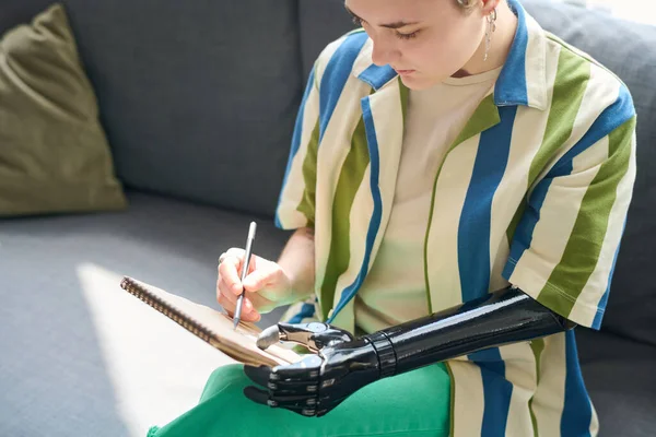 Młoda Kobieta Myoelektryczną Ręką Trzymająca Notatnik Siedząc Kanapie Salonie Rysując — Zdjęcie stockowe
