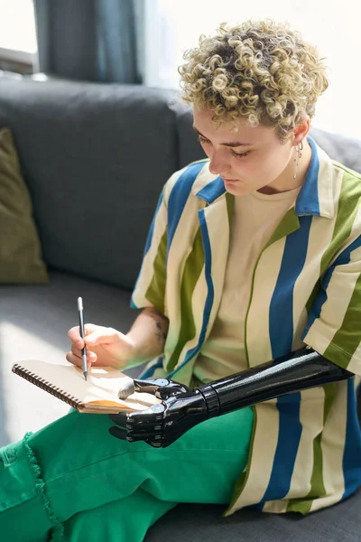 年轻的残疾女性穿着便服坐在客厅舒适的沙发上 用铅笔在笔记本上画着草图 — 图库照片