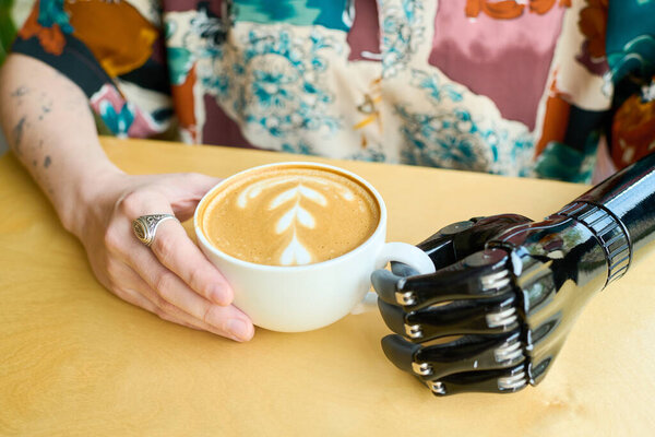 Young contemporary woman with myoelectric arm holding cup of cappuccino decorated with fluffy milk foam in cafe