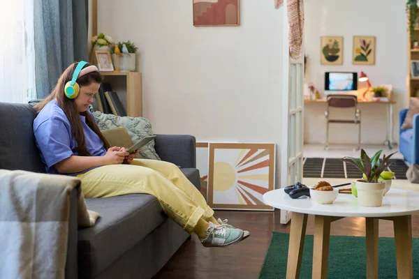女の子とともにダウン症候群でヘッドフォンを見ますタブレット画面で通信中にビデオチャットでソファに座っている間に自宅 — ストック写真