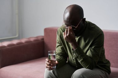 Genç stresli bir adam bir bardak su tutuyor ve psikanalistin ofisinde otururken burun köprüsüne dokunuyor.