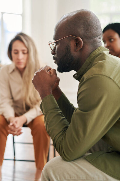 Side view of young stressed man with interlocked hands by his face sharing his problem while attending psychological session