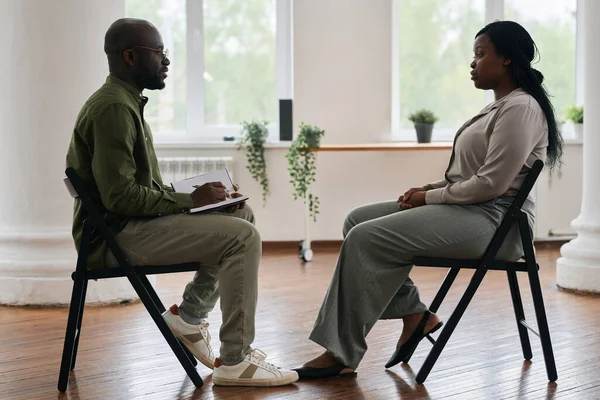 年轻的非洲裔美国男性心理学家 带着笔记本坐在他面前咨询女病人的侧视图 — 图库照片