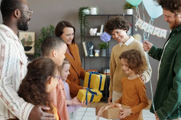 Parents and friends with kids congratulating boy with birthday giving him presents during home party