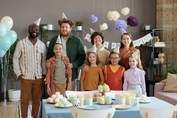 誕生日パーティー中に甘い食べ物とテーブルに立っている子供たちと一緒にカメラで笑顔パーティー帽子の幸せな大人の肖像画 — ストック写真