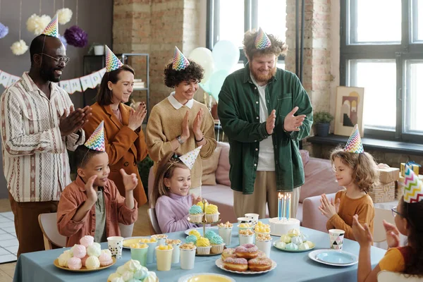 Parents and group of friends with children congratulating little boy with birthday, they sitting at table with birthday cake with candles and clapping hands