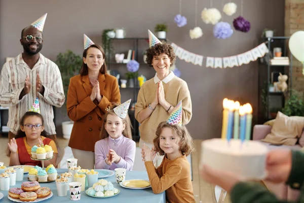 パーティーの帽子の幸せな人々のグループが立っていて デザートとテーブルに座っている子供たちと一緒に誕生日ケーキを待っています — ストック写真