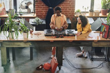 Kulaklık ve günlük kıyafetlerle genç bir çift yeni müzikler yaratırken siyahlar DJ kontrolöründeki sesleri ayarlıyorlar.