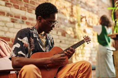Akustik gitarlı genç bir adam dairesinin oturma odasında tuğla duvarın önünde oturan kız arkadaşına karşı oturuyor.