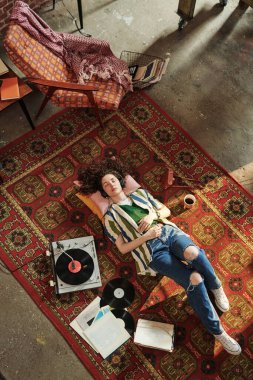 Günlük kıyafetli kız oturma odasında kırmızı halıda vynil disklerle pikabın yanında yatıp kulaklıkla müzik dinliyor.