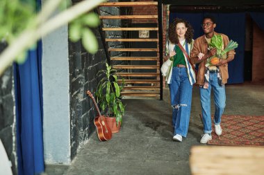 Kültürler arası mutlu çift şık günlük giysiler içinde süpermarketten dönerken tavan arası dairesinin oturma odasında dolaşıyorlar.