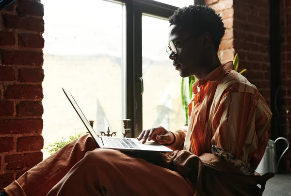 年轻黑人男子靠着窗户坐在阁楼公寓和社交网站上看笔记本电脑屏幕的侧视图 — 图库照片