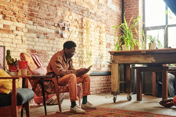年轻的黑人男子穿着便服 用智能手机发短信 或坐在靠墙的扶手椅上视频聊天 — 图库照片