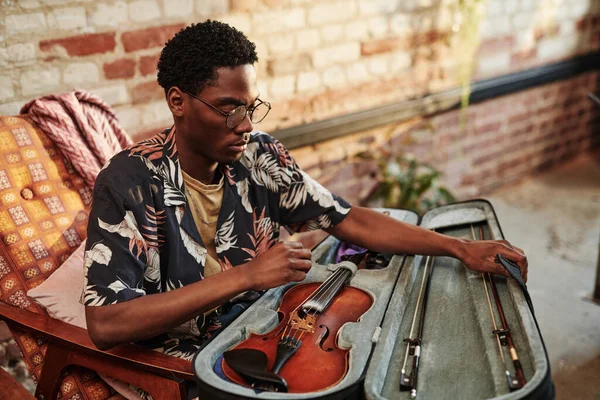 穿着时髦便装的年轻黑人男子在阁楼公寓客厅的扶手椅上坐着时 要穿上带有小提琴和小提琴手的开领拖鞋 — 图库照片