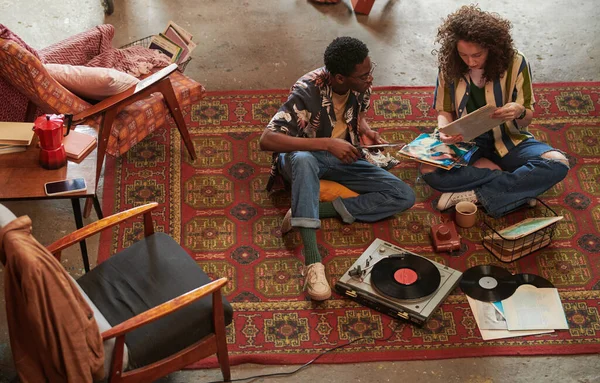 年轻女子穿着时髦的便服坐在旁边的红地毯上 向非洲裔美国男友展示她最喜欢的Vynil盘 — 图库照片