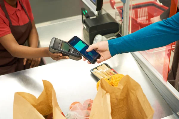 スマートフォンを保有する購入者及びスーパーマーケットにおける食品包装カウンター越しの決済端末を有する販売業者の手 — ストック写真