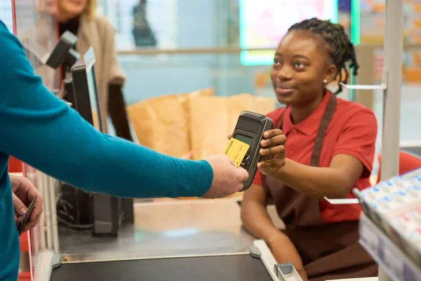 若いアフリカ系アメリカ人女性販売者の支払端末によってそれを保持しながら クレジットカードで選ばれた商品のために支払う現代の買い手 — ストック写真