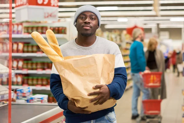 Young African American male buyer carrying paper sack with two fresh wheat baguettes and looking at camera against other customers