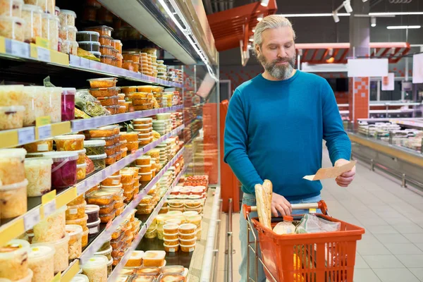 ショッピングリストを見ながらディスプレイと食品とカートを押しながら通路に沿って移動しながら 現代の成熟した男 — ストック写真