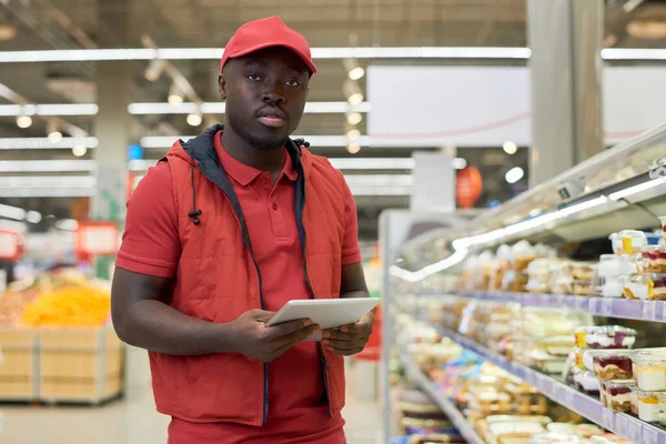 スーパーマーケットでプラスチック容器に詰められたデザートと大きなディスプレイによって立ってタブレットと深刻な若い黒人男性 — ストック写真