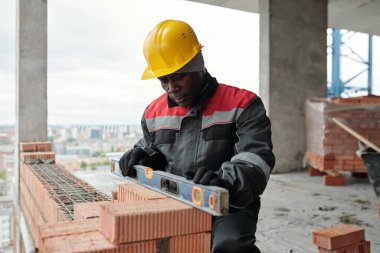 İş giysisi ve koruyucu miğfer konusunda olgun müteahhit, duvarın düz olup olmadığını kontrol ederken aynı zamanda tuğla tabakasının üzerinde el aletini tutuyor.