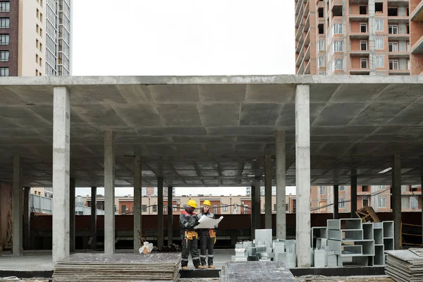 未完成のコンクリート構造物の中に立っている間 建設現場で新しい建物のスケッチを議論する作業服の2人の異人種間の建築家 — ストック写真