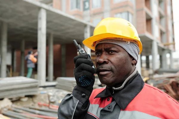 Confident African American male engineer in uniform and protective helmet speaking to subordinates in walkie-talkie during work