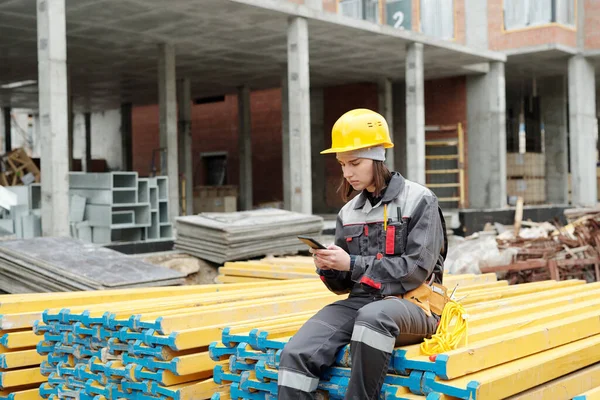 穿着工作服的年轻女性坐在一堆黄色的木板上 一边用智能手机发短信 一边在建筑工地下班休息 — 图库照片