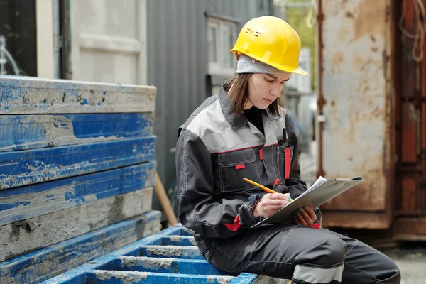 年轻而严肃的女工程师坐在建筑材料上 戴着防护头盔 穿着工作服 在剪贴板上画草图 — 图库照片
