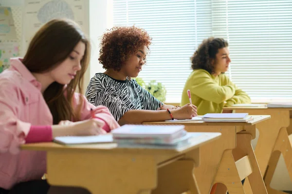 アフリカ系アメリカ人の深刻な高校生は クラスメートの間で机に座ってノートを作りながら授業で仕事に集中します — ストック写真