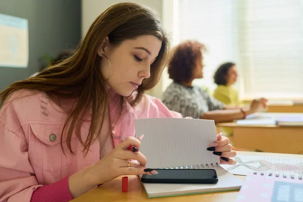 十代の女の子でピンクデニムジャケットテキストでスマートフォンでレッスン後にページのコピーブックしばらく座っていますに対して彼女のクラスメート — ストック写真