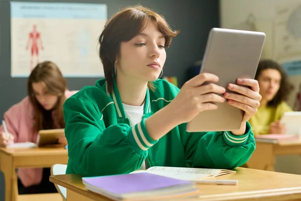 机に座ってオンライン情報を見ている間 彼女の顔の前にタブレットを保持している高校生の深刻な女性 — ストック写真