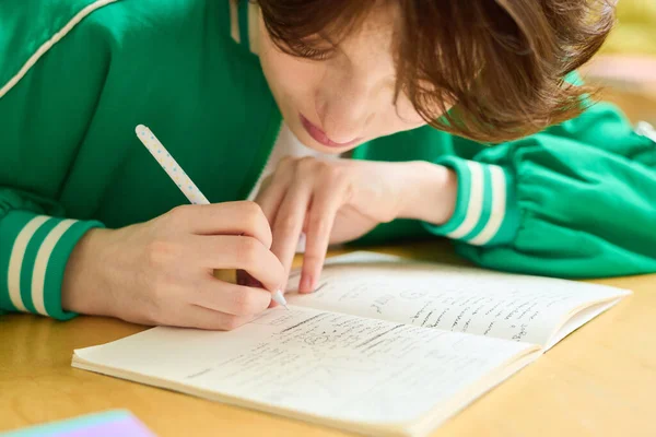 穿着绿色夹克的少女弯腰坐在课桌前 一边在抄袭本上记笔记 一边在课上逐节课 — 图库照片