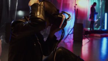 Kafası tıraşlı, deri elbiseli, VR gözlükleri takan, stüdyoda oturan, duvarlarında plastik film ve neon ışıklarıyla beyaz bir kadının yan görüntüsü.