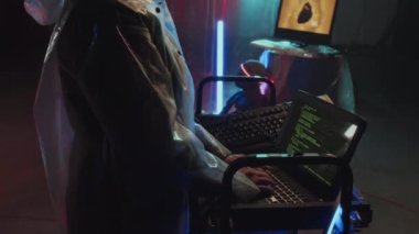Yağmurluk giyen, taşınabilir bilgisayarda kod yazan, duvarlarda plastik film ve neon ışıklarla saklanan genç bir kadının yan görüntüsü.