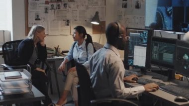Orta uzunluktaki genç siyahi adam bilgisayar kullanıyor, federal suç veri tabanını araştırıyor, iki kadın dedektif ofiste oturuyor ve konuşuyor.