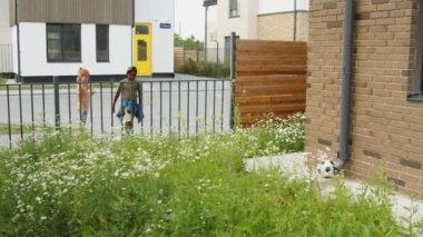 Modern yerleşim yerlerinin yakınındaki sokakta taş-kağıt-makas oynayan iki farklı çocuğun geniş açılı görüntüsü. Sonra siyah çocuk çitten bahçeye tırmanıyor, futbol topunu alıyor ve iki arkadaşı da kaçıyor.