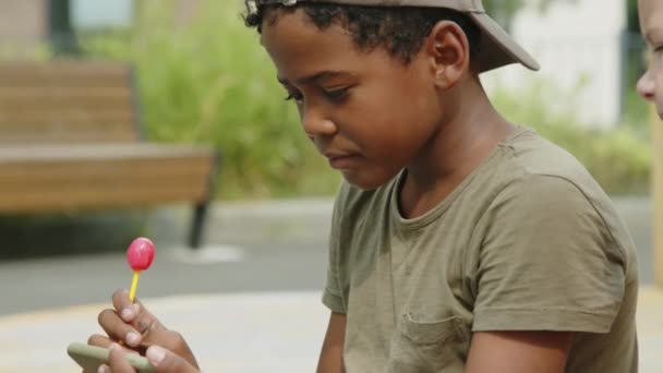 티셔츠를 모자를 벤치에 아프리카 미국인 소년의 스마트폰에서 만화를 친구가 너머로 — 비디오