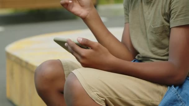 夏休みに公園のベンチで凍えているアフリカ系アメリカ人と白人の子供2人が スマートフォンを使いおしゃべりをしながらロリポップを食べ — ストック動画