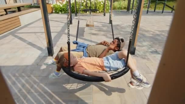 2人のアフリカ系アメリカ人と白人の少年が夏の晴れた日に遊び場で屋外でリラックスし 大きな巣のスイングで横になり 前後にロッキング — ストック動画
