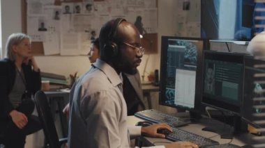 Bilgisayarı kullanan genç siyahi bir adamın federal suç veri tabanını ararken yan görüntüsü, ofiste oturan ve konuşan iki kadın dedektif.