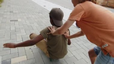 Yazın dışarıda eğlenen iki çocuğun orta boy arka plan çekimi kaykayda oturan Afrikalı Amerikalı bir çocuk ve beyaz bir arkadaş onu kaldırımdan aşağı itiyor.