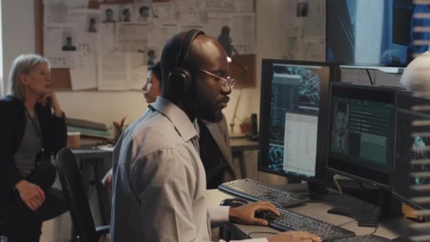 コンピュータを使って黒人青年を横目に連邦犯罪データベースを探してる2人の女性刑事がオフィスに座って話をしてる — ストック動画