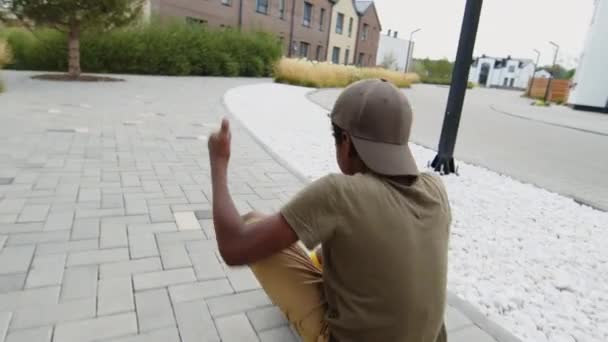 野球帽をかぶったアフリカ系アメリカ人の少年の中長い追跡ショットスケートボードに座って叫んで笑って そして白人の友人は彼を舗装された通りを押し下げる — ストック動画