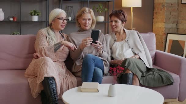 Oturma Odasındaki Mor Koltukta Oturan Olgun Kadın Akıllı Telefondaki Fotoğraflara — Stok video
