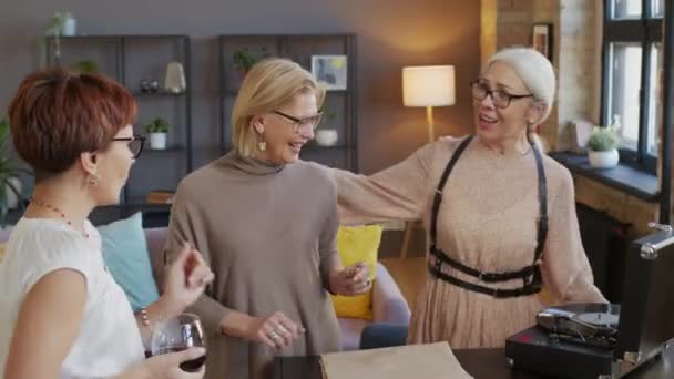 3人の現代の大人の女性が赤ワインを飲み レコードプレーヤーから演奏する音楽に合わせて踊る 現代のロフトスタイルのアパートでホームパーティーを持っている — ストック動画
