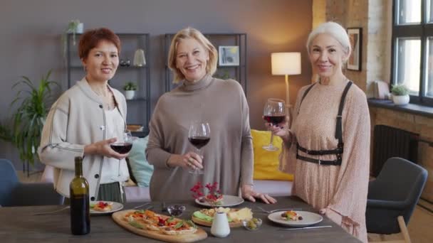 赤ワインのグラスを持っている3人の幸せな成熟した女性の肖像画とカメラで笑顔 現代のロフトスタイルのアパートでディナーテーブルのそばに立って — ストック動画
